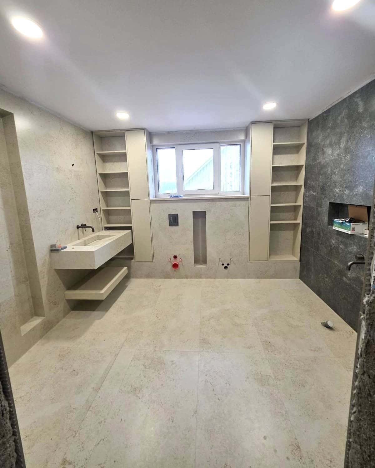 Baie cu plăci cu aspect de piatră bej, rafturi încastrate lângă fereastră și lavoar suspendat