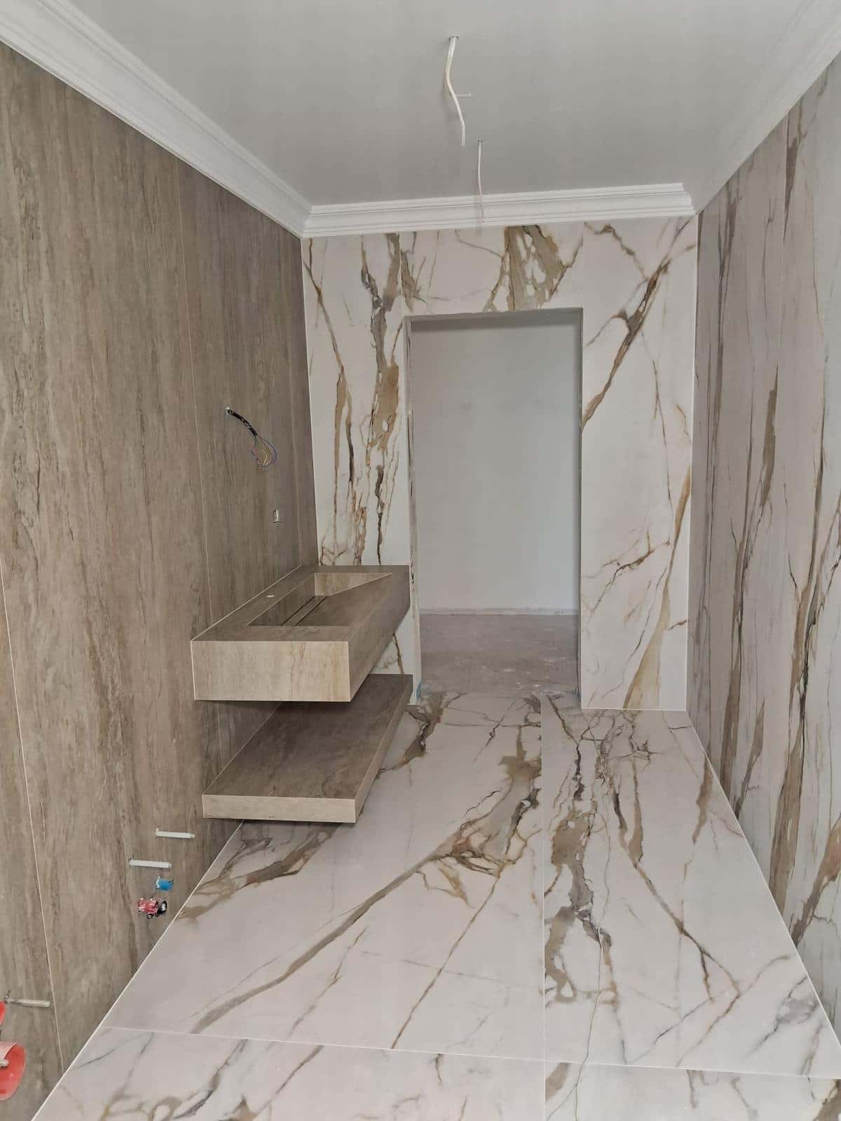 Baie îngustă cu un perete cu aspect de travertin, lavoar placat suspendat și plăci albe cu vinișoare aurii