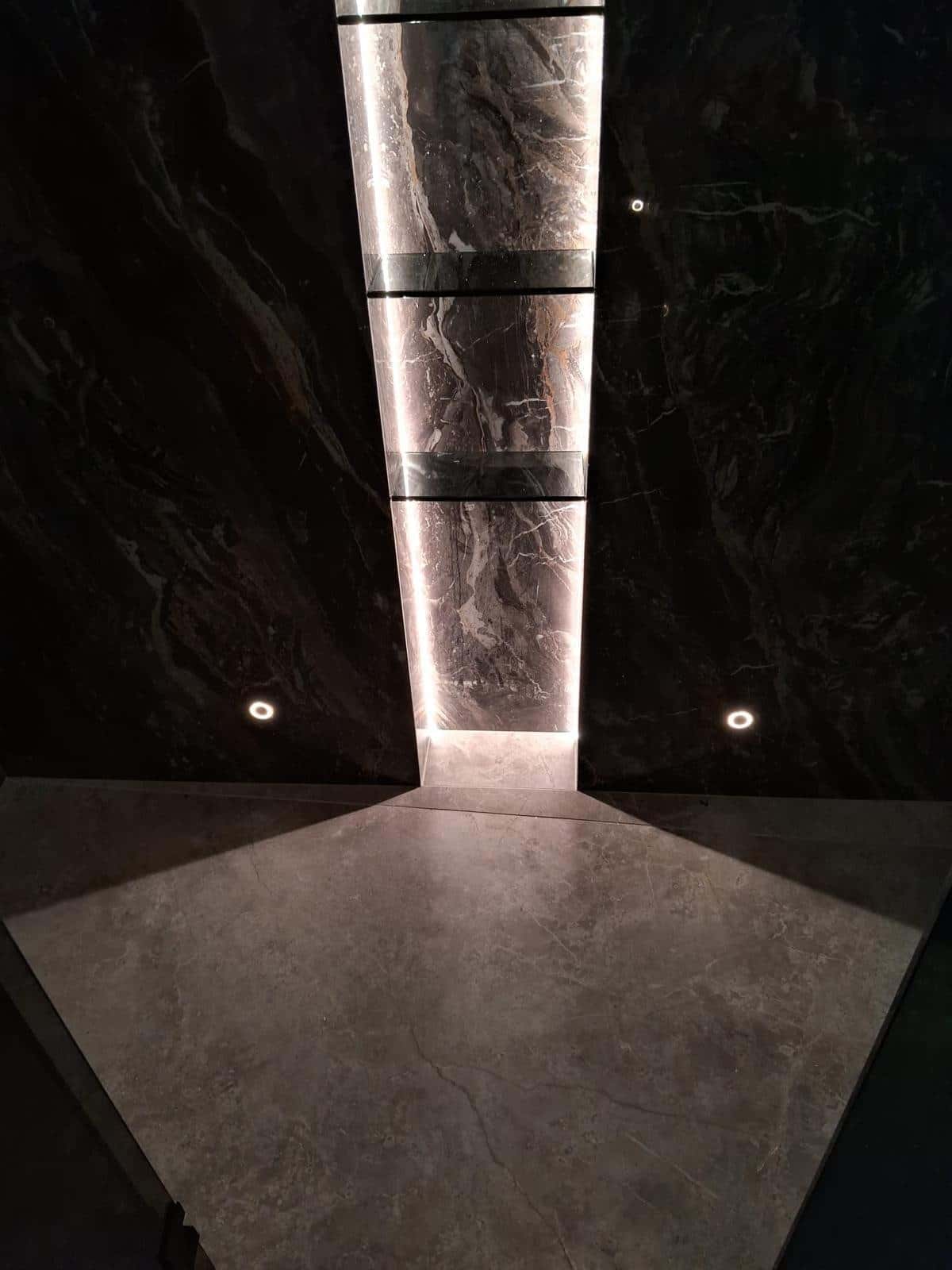 Pereți cu aspect de marmură neagră și nișă verticală luminată, cu rafturi de sticlă