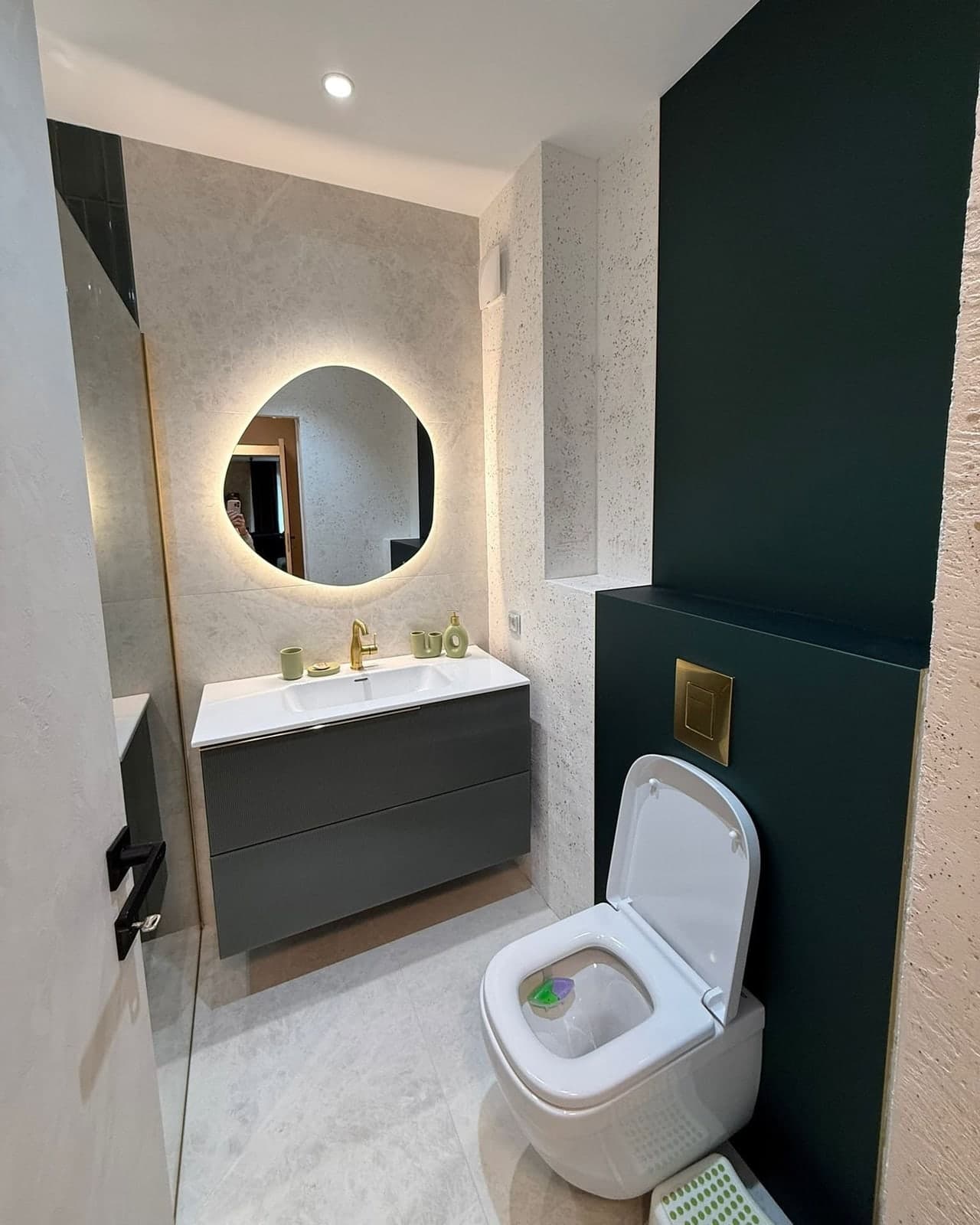 Baie cu oglindă organică luminată, mobilier gri cu baterie aurie și perete verde închis